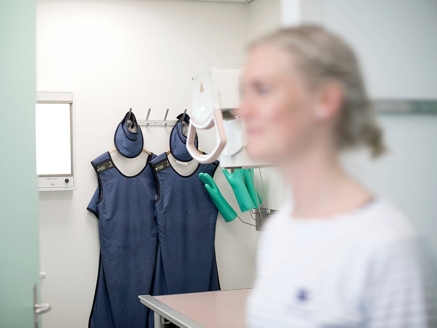 Eine Frau ist unscharf im Vordergrund zu erkennen und scheint sich zu unterhalten. Im Hintergrund hängt OP-Bekleidung an der Wand.
