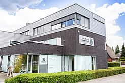 Das Haus, in dem sich die Praxis befindet, ist modern und hat große Fenster. Drum herum ist eine grüne Hecke.