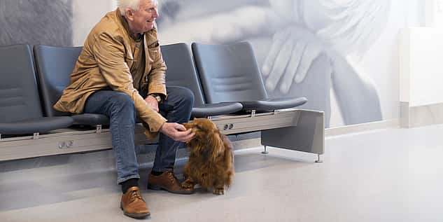 Ein älterer Herr sitzt mit seinem Dackel im Wartezimmer. Er lacht und streichelt ihn dabei.