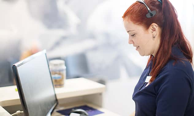 Eine Frau mit Headset sitzt vor einem Computerbildschirm. Sie lächelt, während sie darauf schaut.