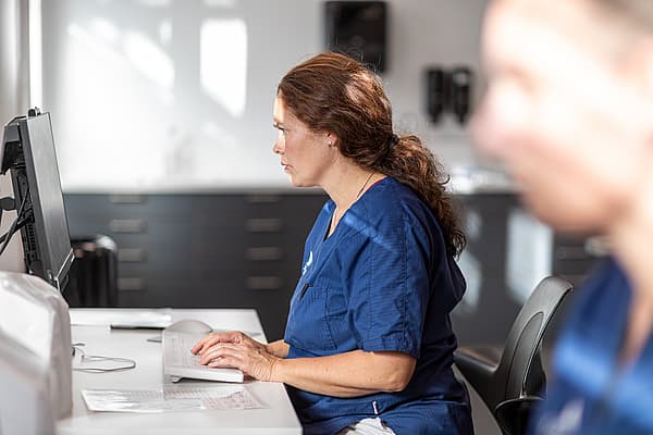 Eine Person in blauem Kittel sitzt an einem Schreibtisch und bedient einen Computer.