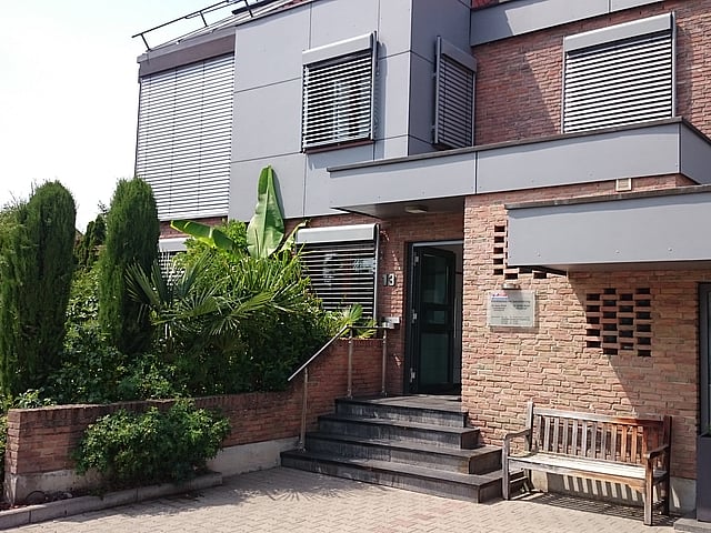 Der Eingang der Praxis ist in einem Backsteinhaus, mit modernen grauen Elementen. Links befinden sich viele Pflanzen und rechts steht eine Bank.